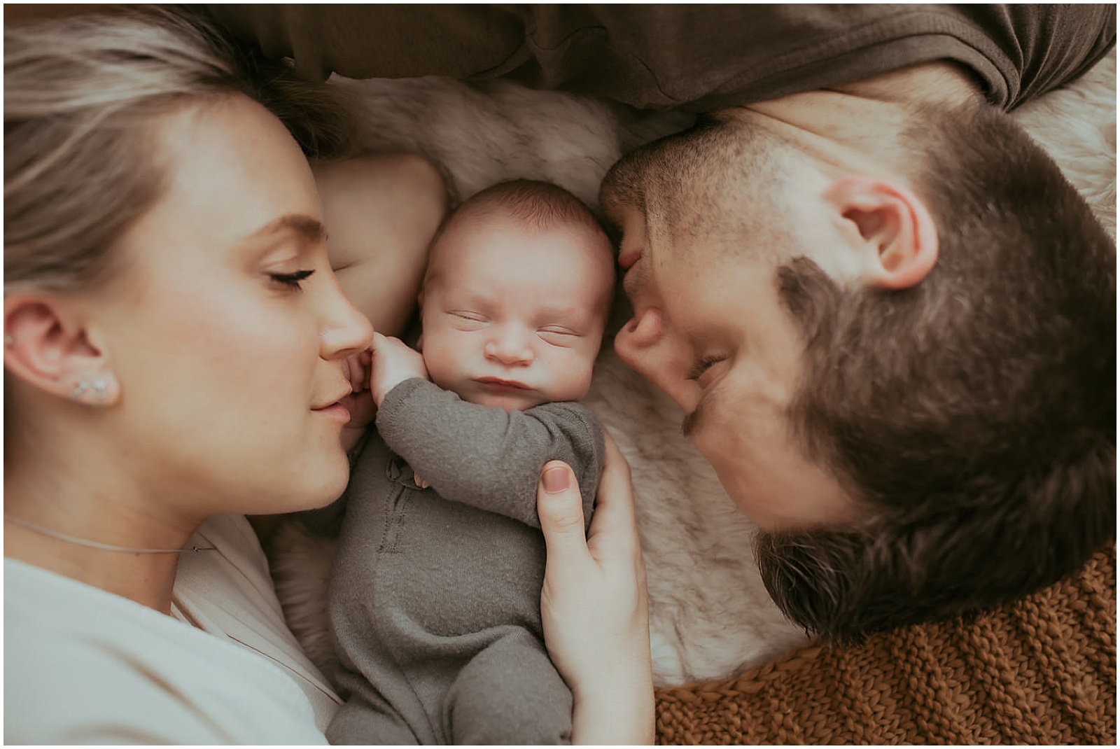 A mother and father lay on either side of a newborn while a photographer takes their picture.