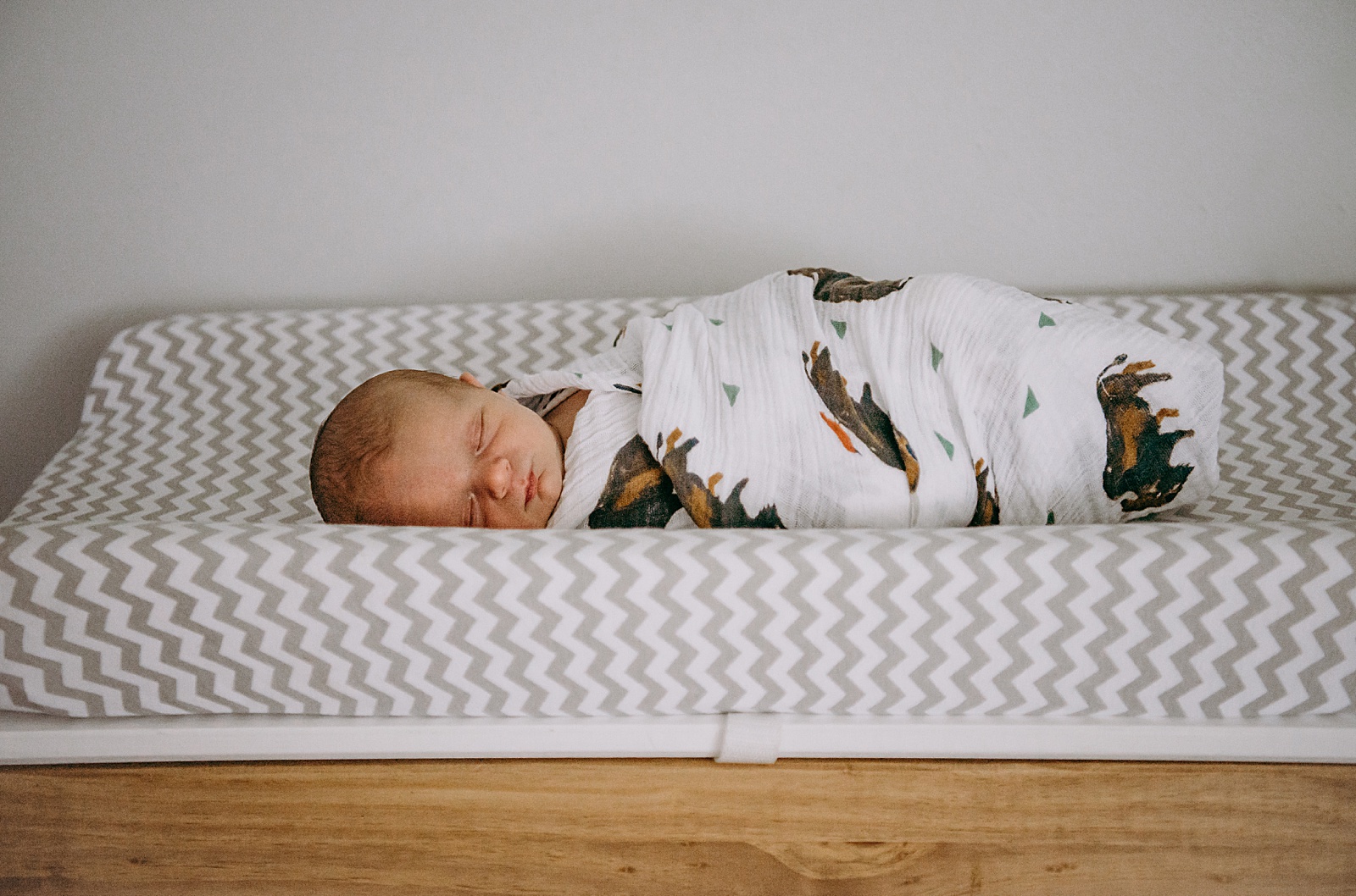 A swaddled newborn lays on a changing pad on top of a dresser.