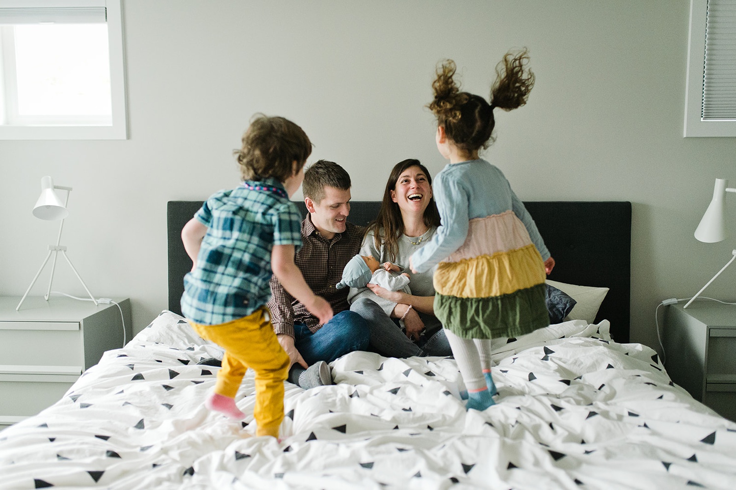 Two parents laugh and hold their baby while their toddlers jump on a bed in lifestyle family photo.