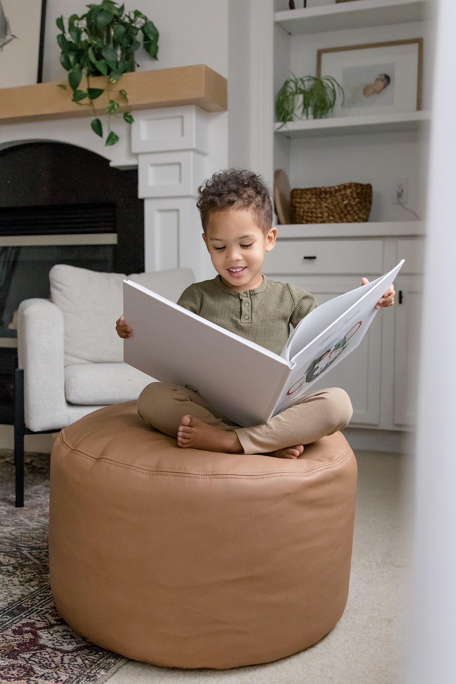 A little boy sits on an ottoman smiling while he reads a baby book.