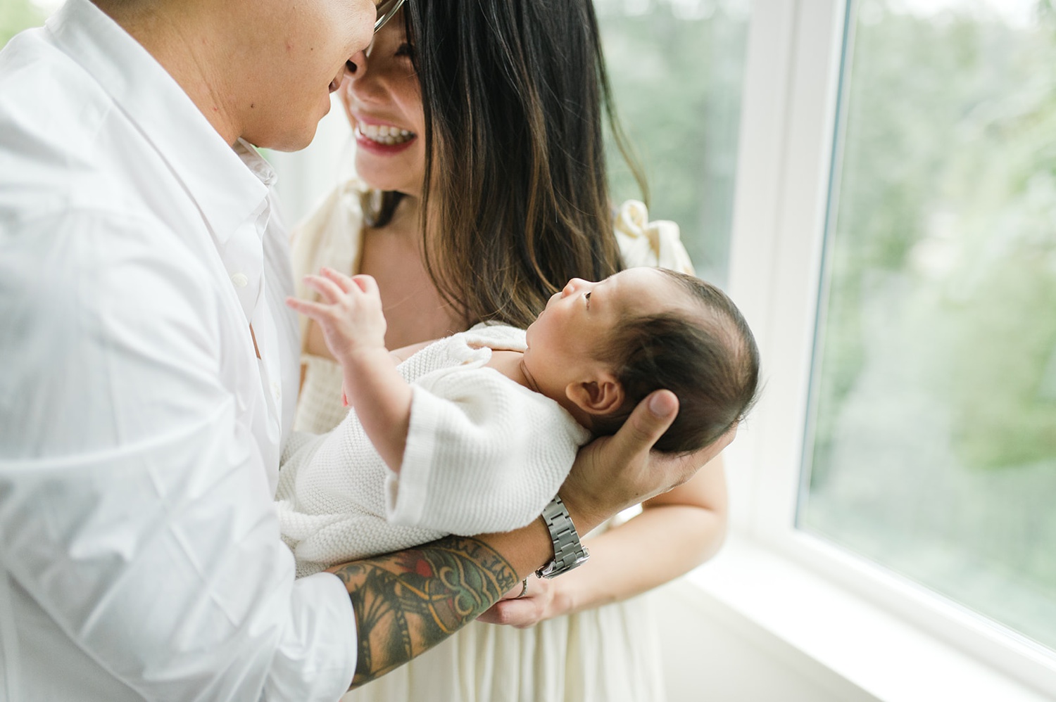 A mother and father smile and hold their newborn beside a window.