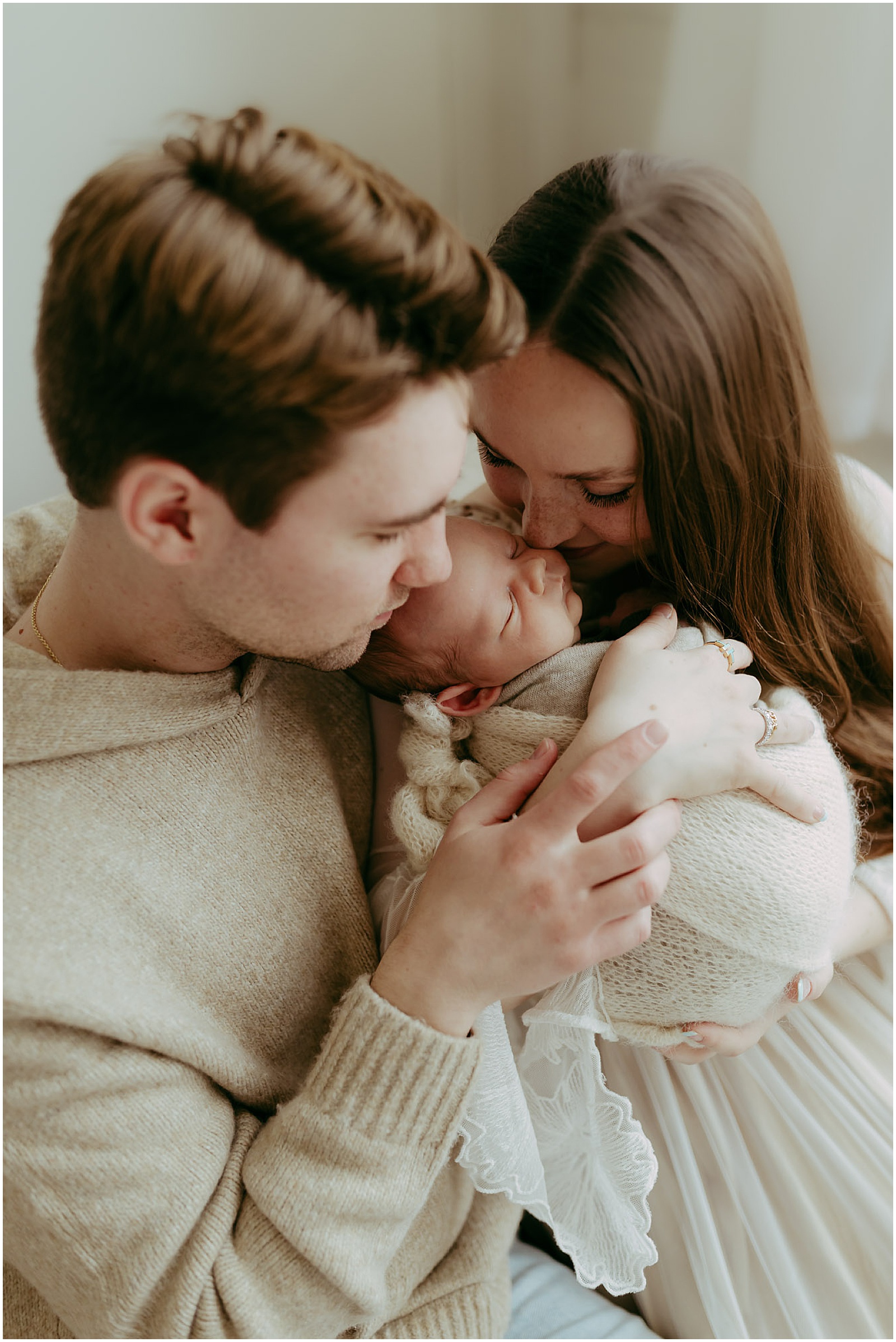 A mother and father hold a baby while a newborn photographer takes their picture.