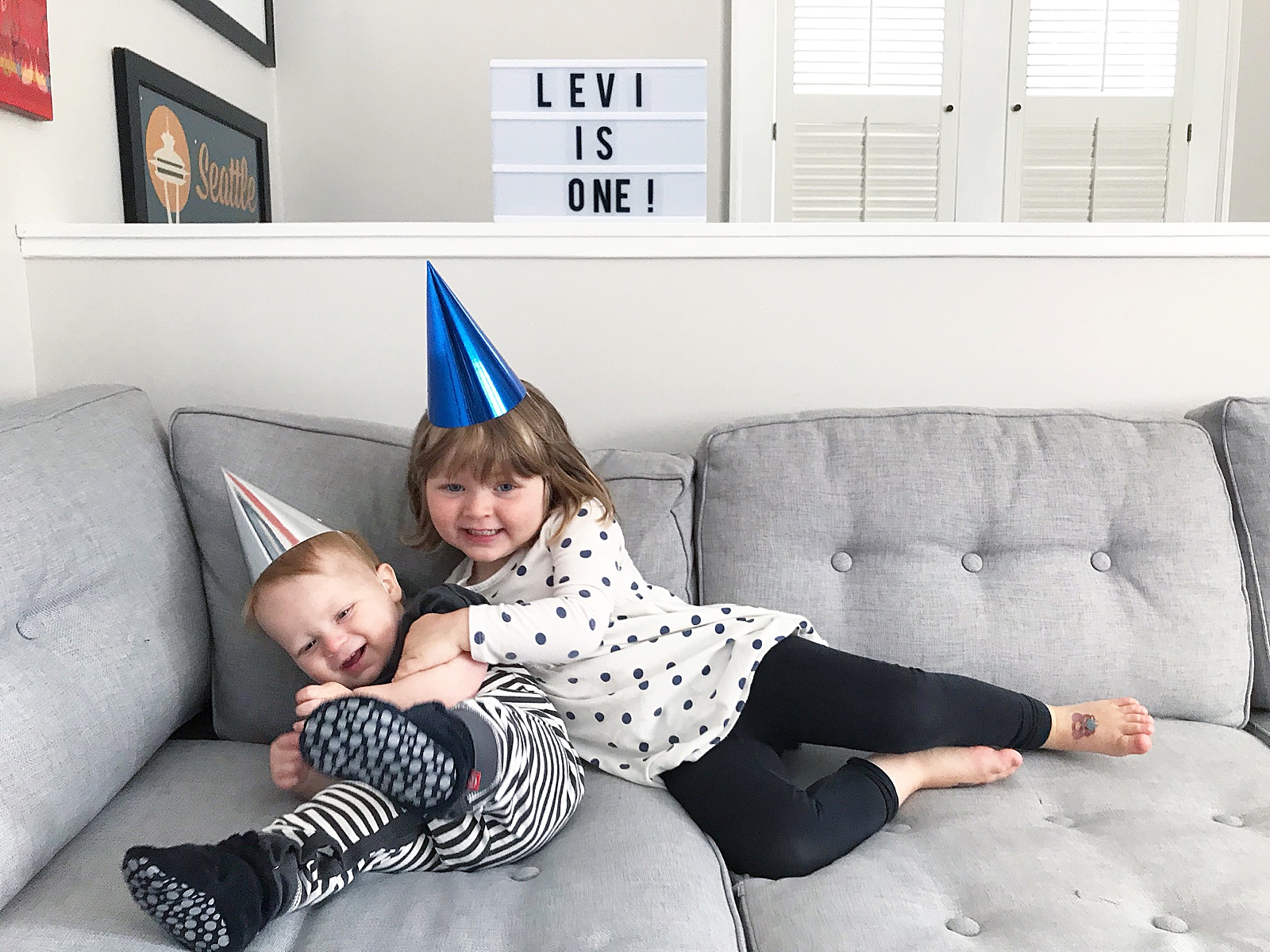 A little girl leans across a gray couch to hug her baby brother.