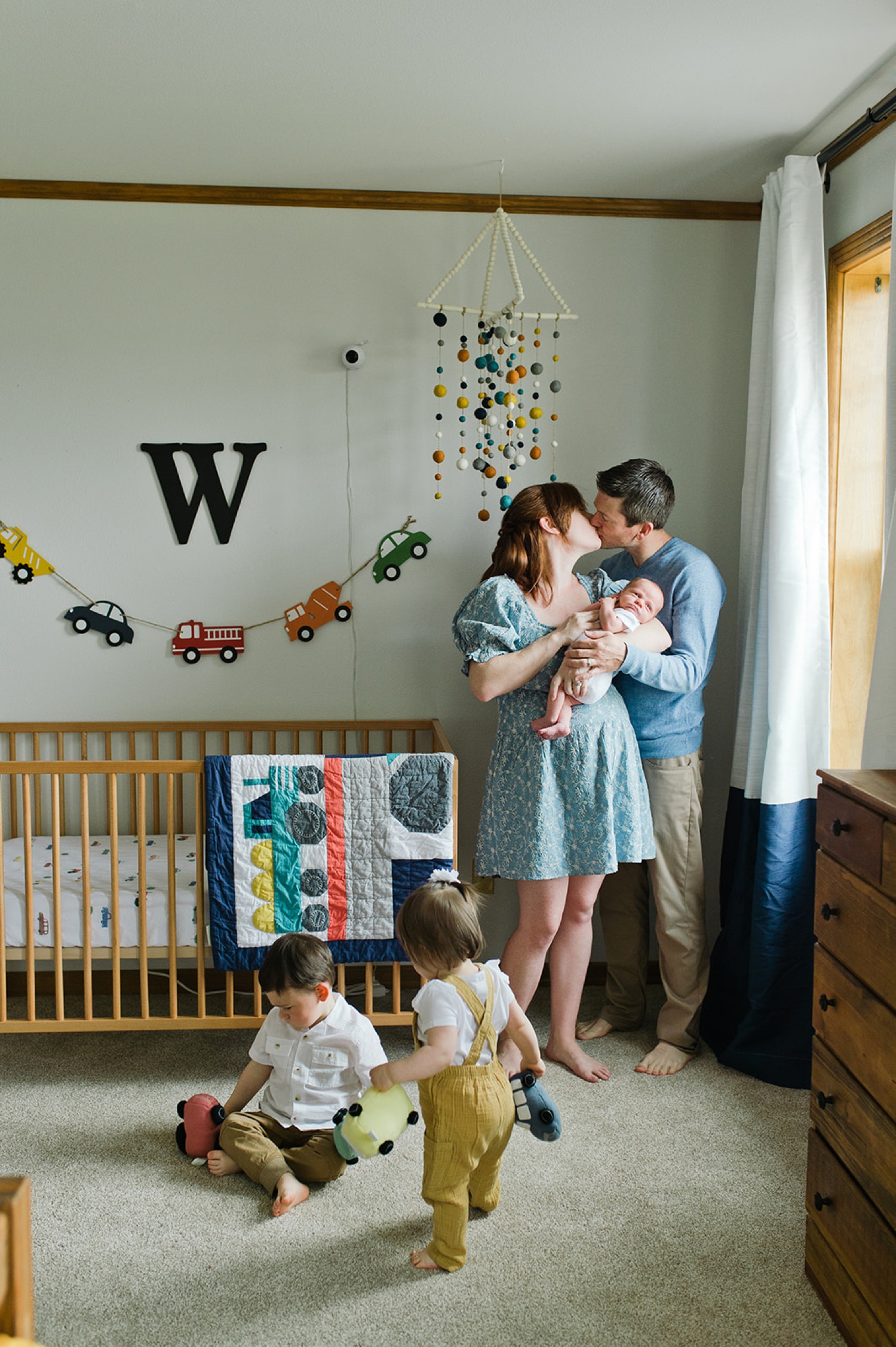 A couple kisses and holds their newborn baby while their twins play on the floor in a newborn photoshoot.