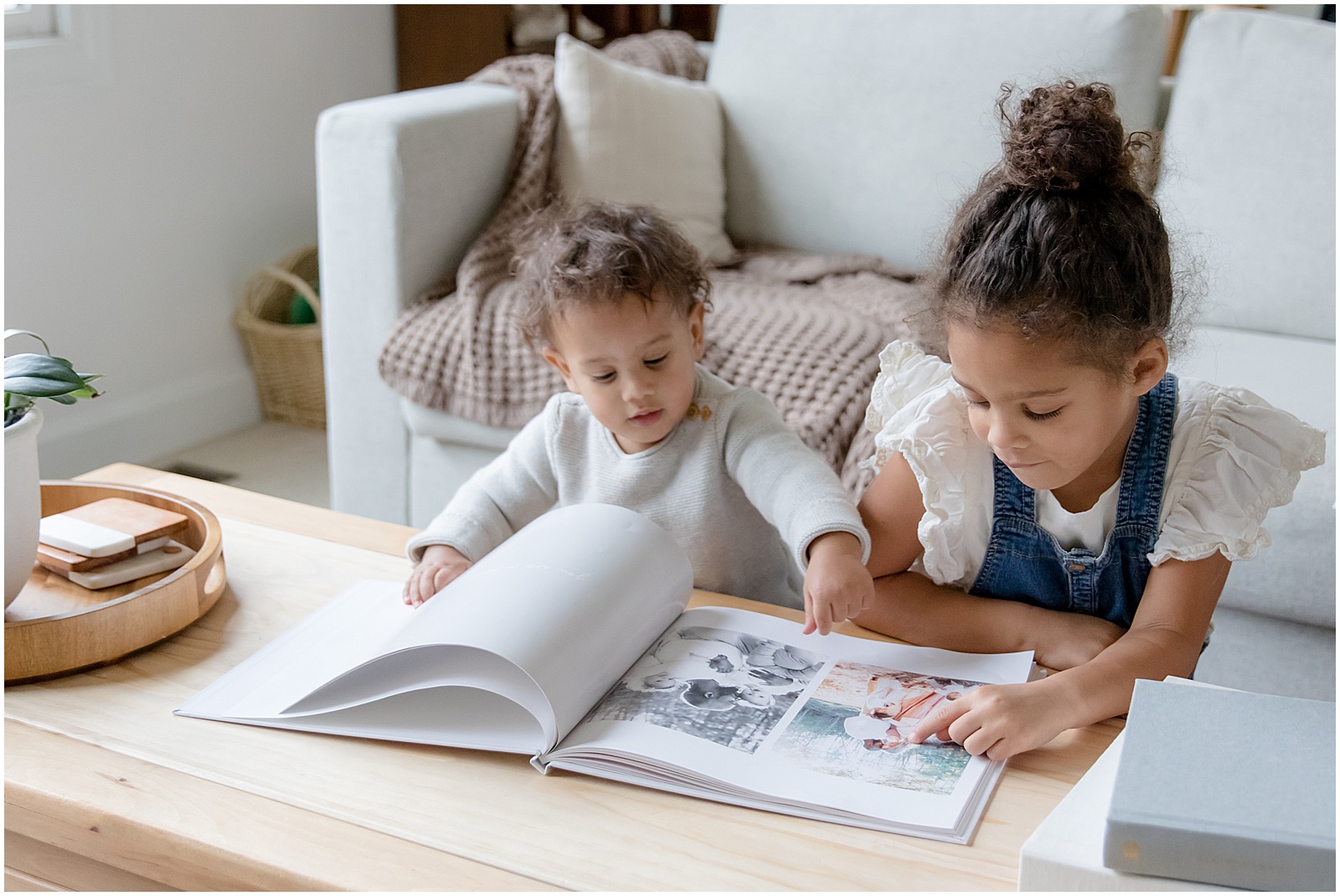 Siblings sit at a coffee table looking at a baby book made with a memory book app.