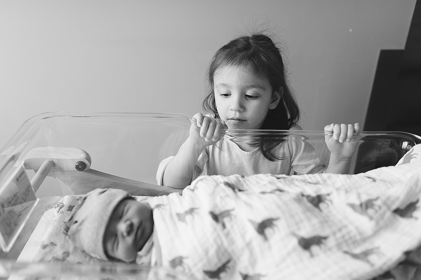 A little girl looks over the edge of a hospital bassinet at a swaddled newborn.