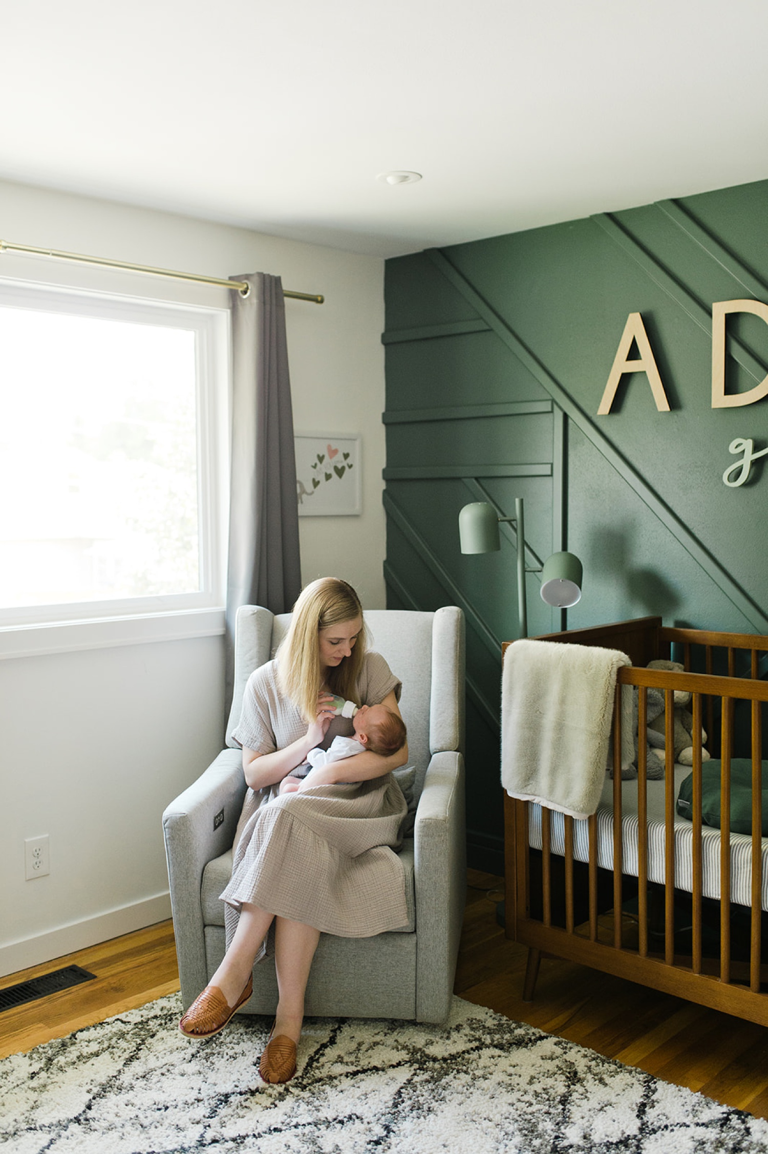 A woman sits with a newborn in a chair in the corner of a nursery during a newborn photoshoot.