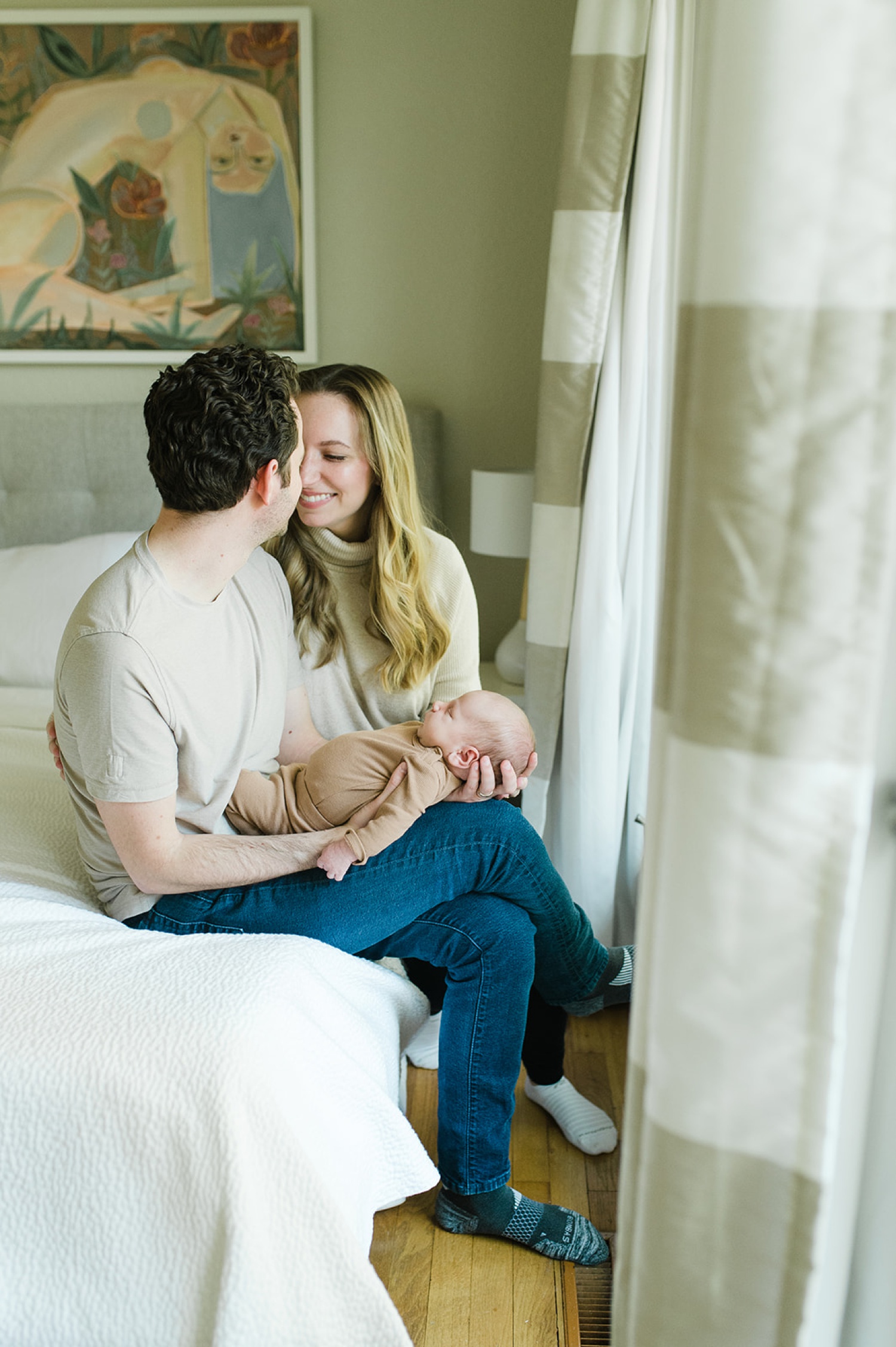 A couple sits on a bed holding their baby in a newborn photoshoot at home.