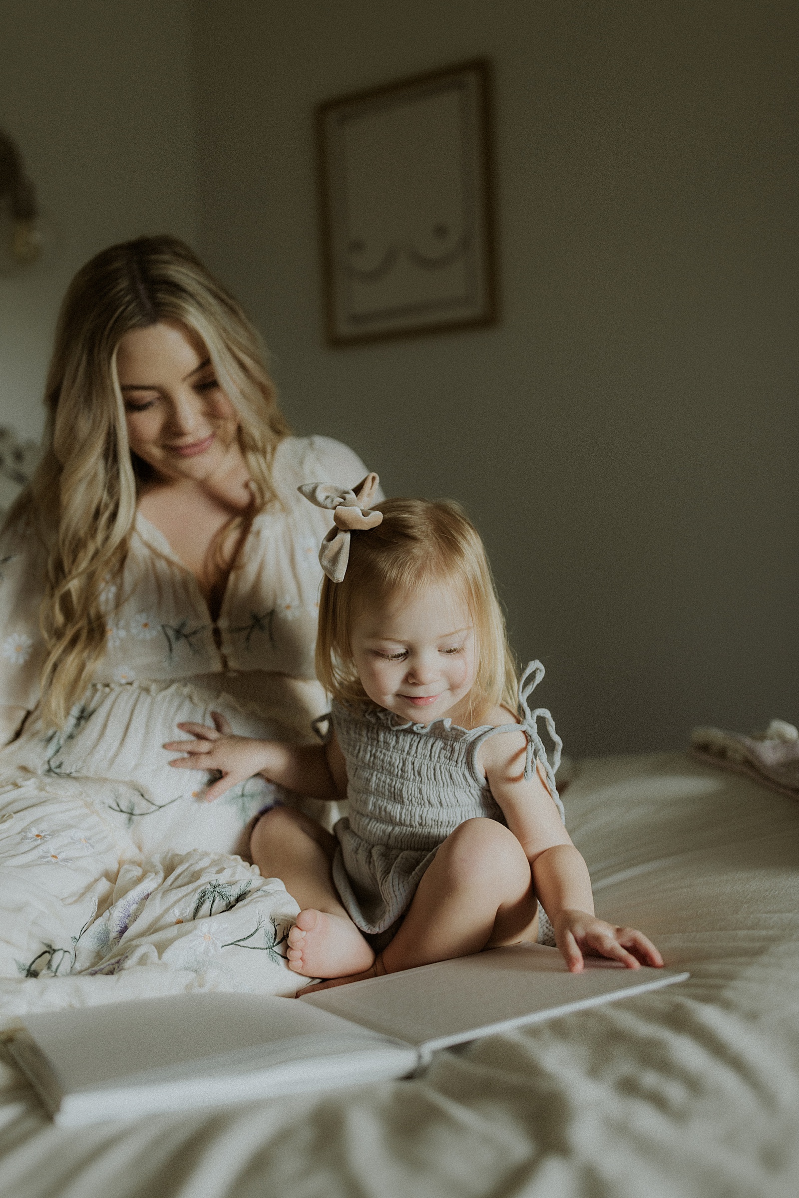 A woman sits on a bed flipping through a pregnancy memory book with her toddler.