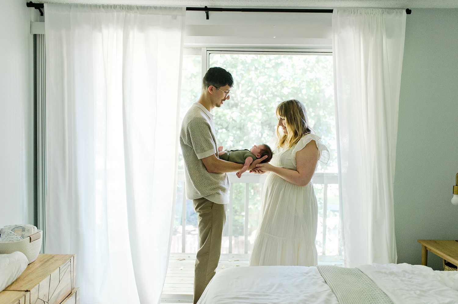 A man and woman hold their newborn in front of an open window during a newborn photoshoot.