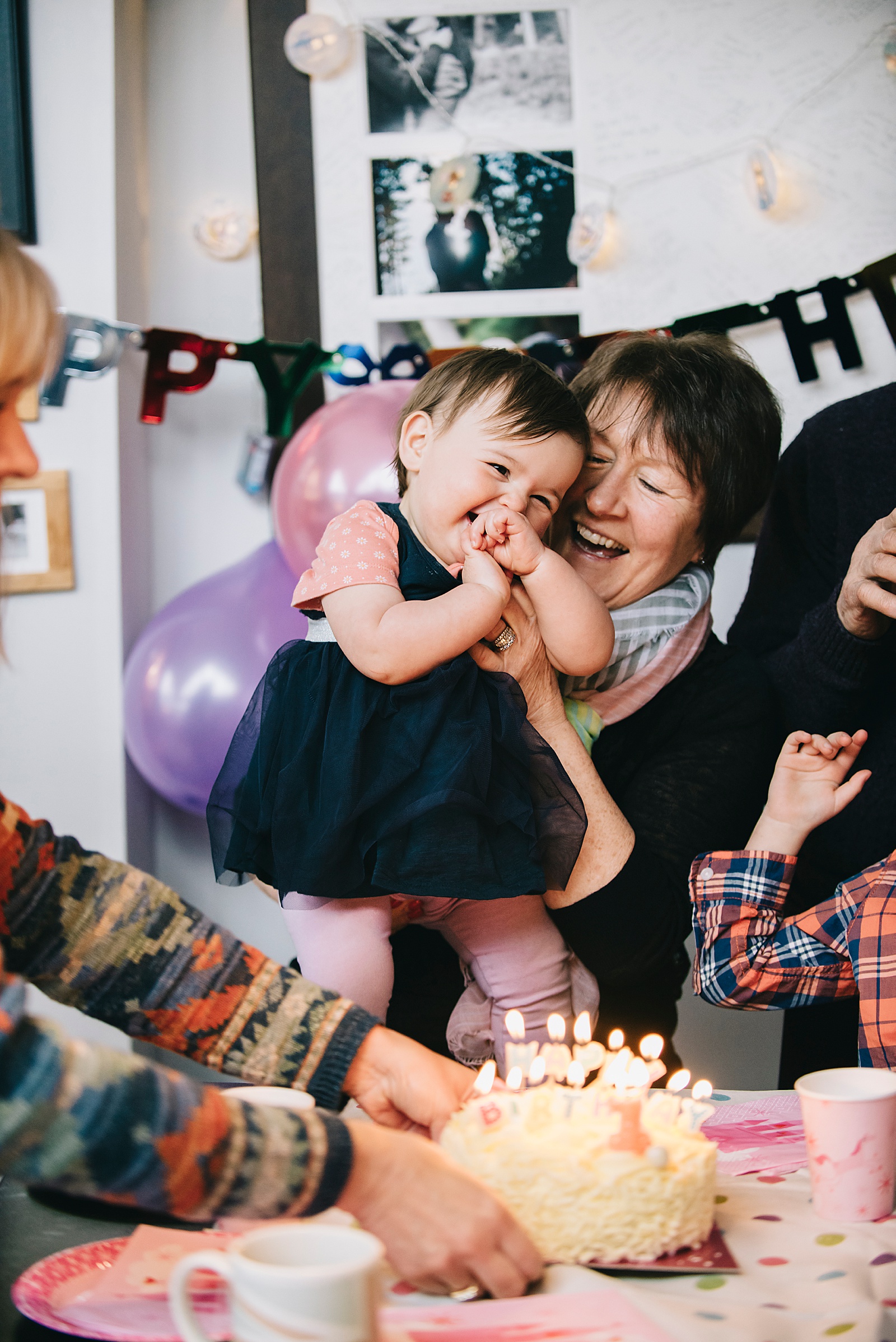 A grandmother sits at a table hugging a baby as someone sets a cake on the table.