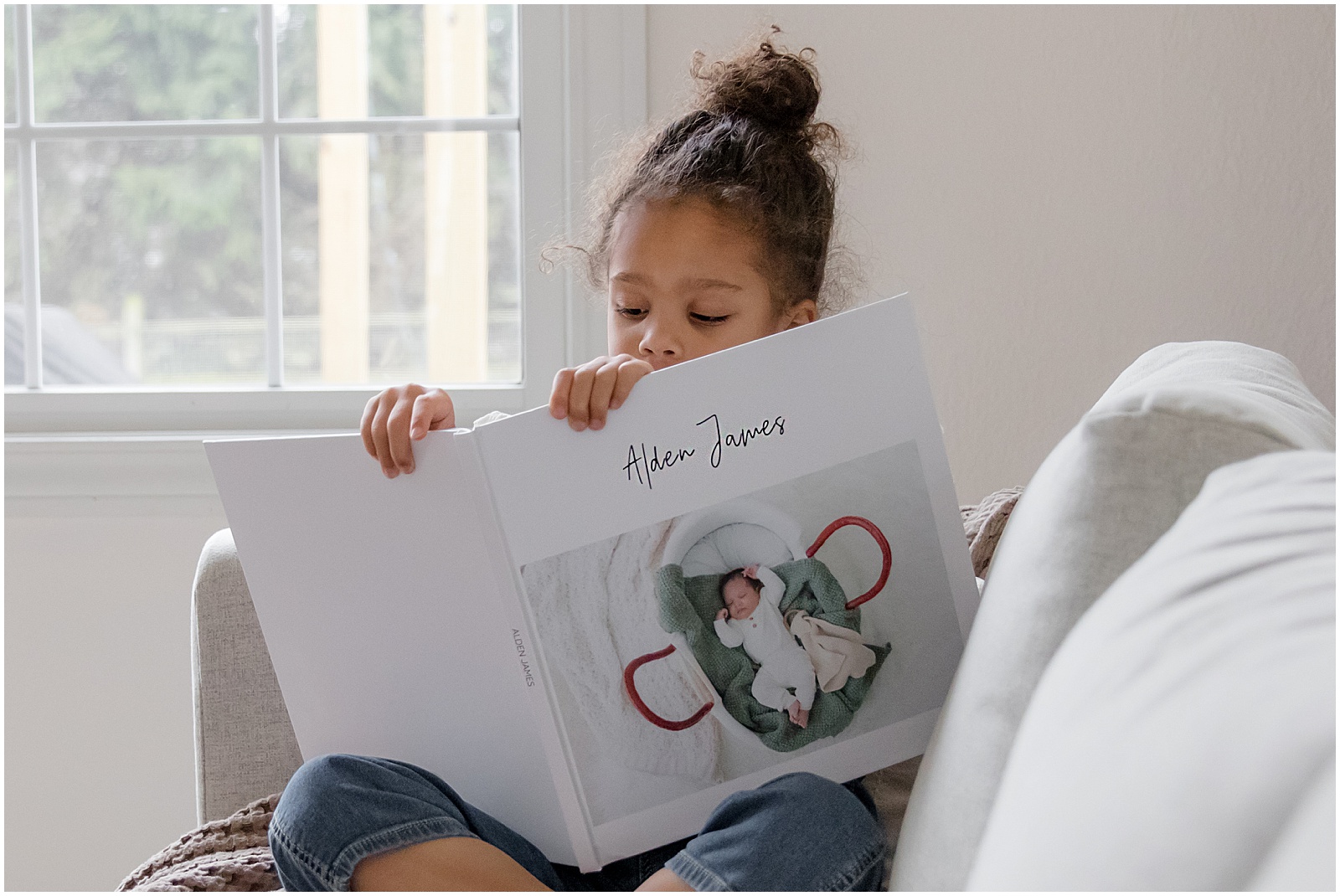 A toddler looks through his baby book while sitting beside a window.