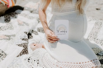 A pregnant woman sits on a crocheted blanket holding a pregnancy card beside her baby bump.