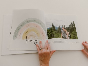 A woman opens a baby book to a family photo in a forest.