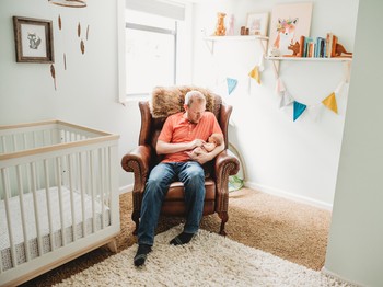 A father holds a newborn in an armchair in the corner of a minimalist nursery.