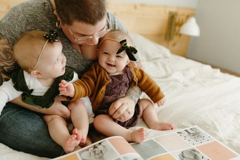 A mother sits with her twins in her lap flipping the pages of a modern twin baby book.